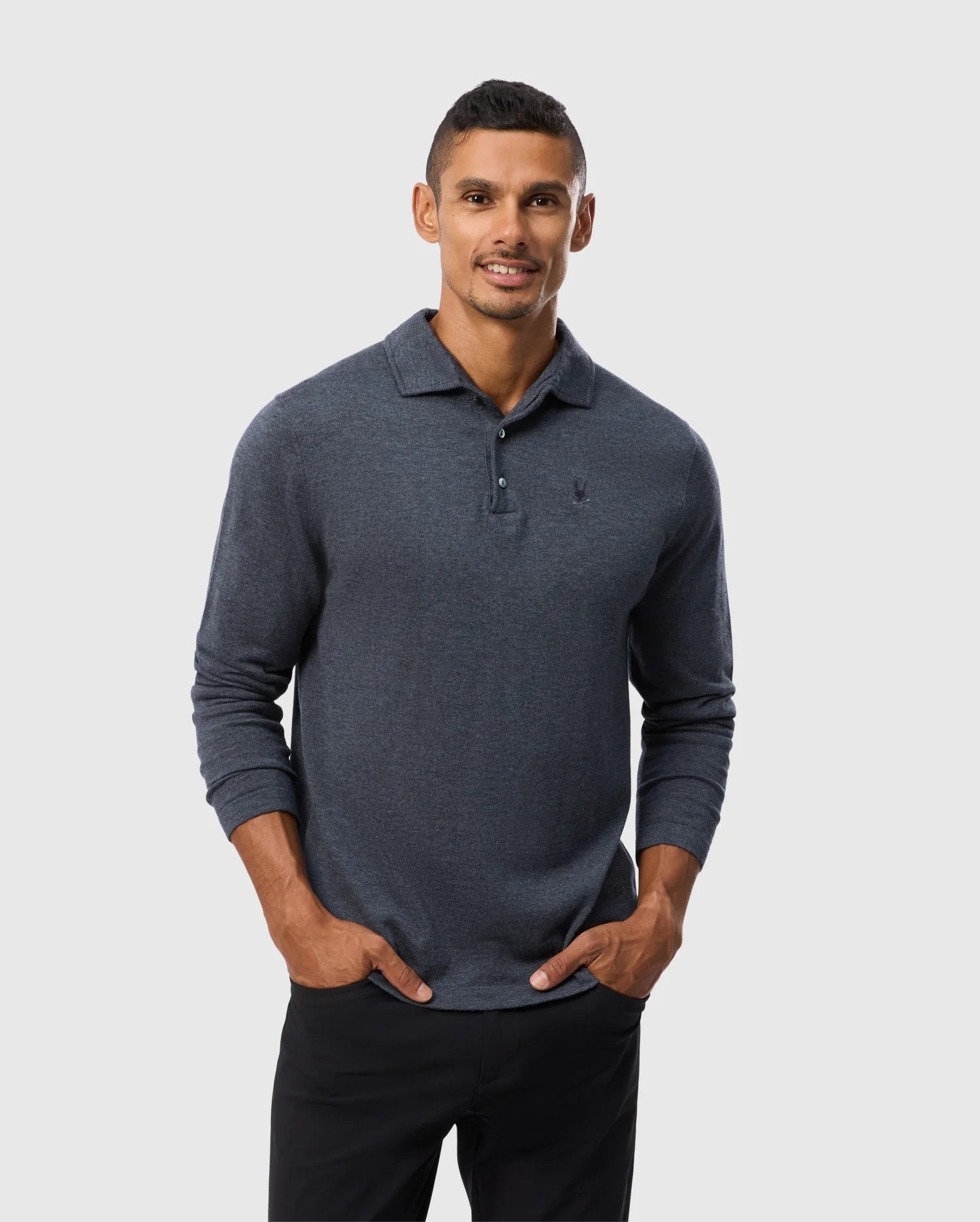 A man is standing against a light grey background, dressed in the dark grey MENS BRADSHAW POLO SWEATER - B6E835D200 from Psycho Bunny. The premium merino blend shirt features long sleeves, a collared design, and a partially buttoned front. He has his hands in his black pants pockets and is smiling at the camera.