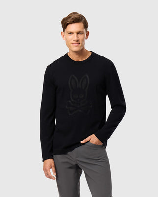 A man with short hair is wearing the MENS CORY CHAINSTITCH COTTON SWEATER (B6E893D200) by Psycho Bunny, which features a subtle bunny and crossbones design on the chest. He is also dressed in grey pants, standing against a plain light grey background with a relaxed demeanor and one hand in his pocket.