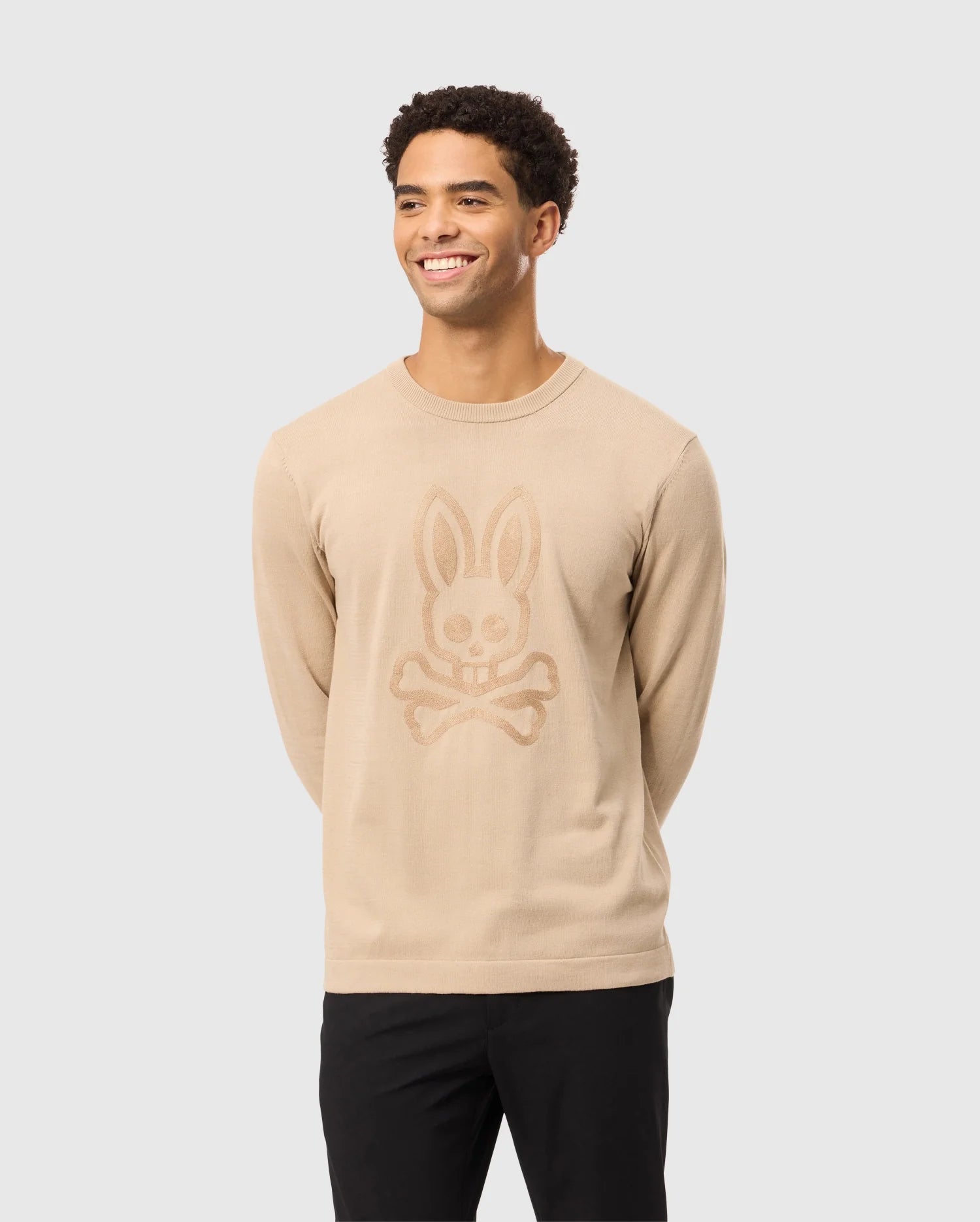 A man with short, curly hair is smiling and wearing a Psycho Bunny MENS CORY CHAINSTITCH COTTON SWEATER - B6E893D200 that features a stylized bunny skull and crossbones design in chainstitch embroidery on the front. He is standing against a plain light gray background, with his hands behind his back, dressed in black pants.