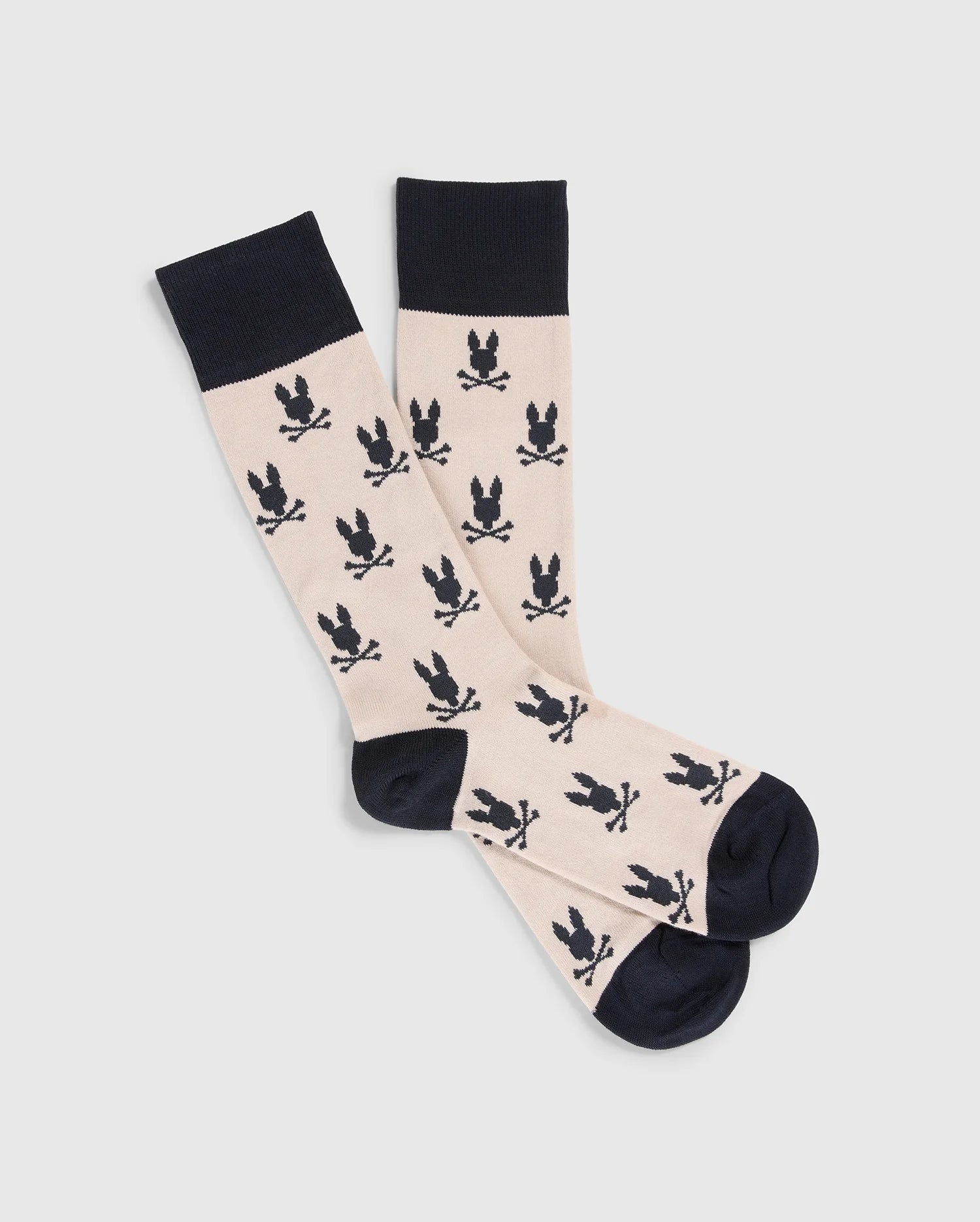 A pair of MENS DRESS SOCKS - B6F750C200 by Psycho Bunny, featuring a beige base adorned with black skull and crossbones emblems. These luxurious socks are crafted from Peruvian Pima cotton, complete with black cuffs, heels, and toes, and are displayed flat against a plain background.