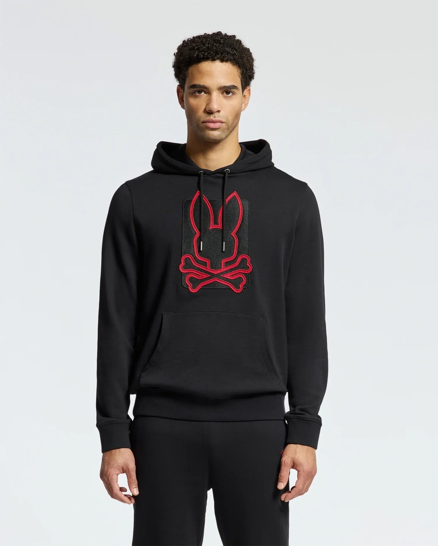 A person wearing the MENS PIERCE ESSENTIAL FRENCH TERRY HOODIE by Psycho Bunny, featuring a red embroidered outline of a stylized bunny and crossbones on the front, crafted from high-quality cotton, stands against a light gray background.