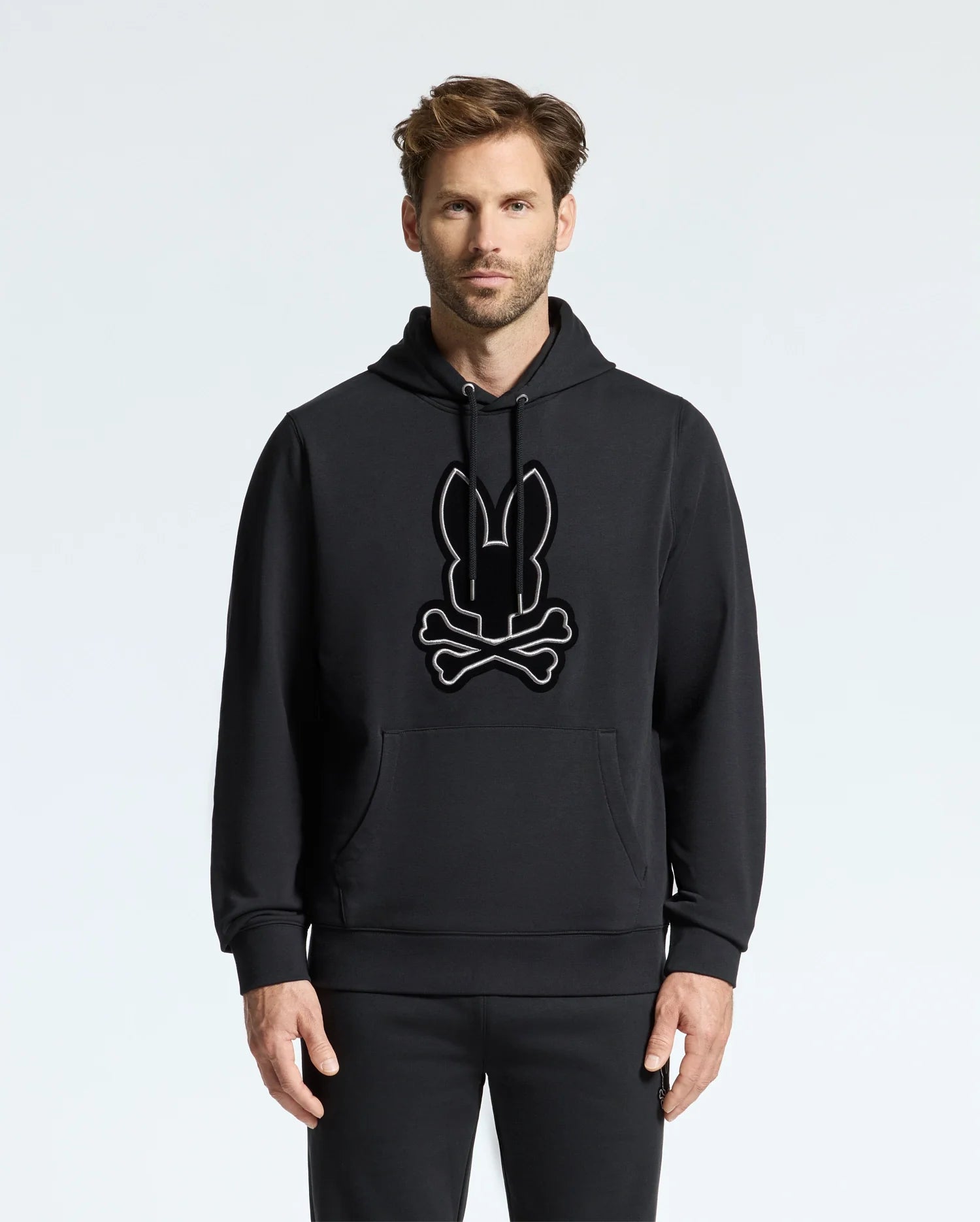 A person in a comfortable Psycho Bunny MENS ALEXANDER HOODIE (B6H154E200) featuring a large rabbit and crossbones graphic stands against a plain, light background.