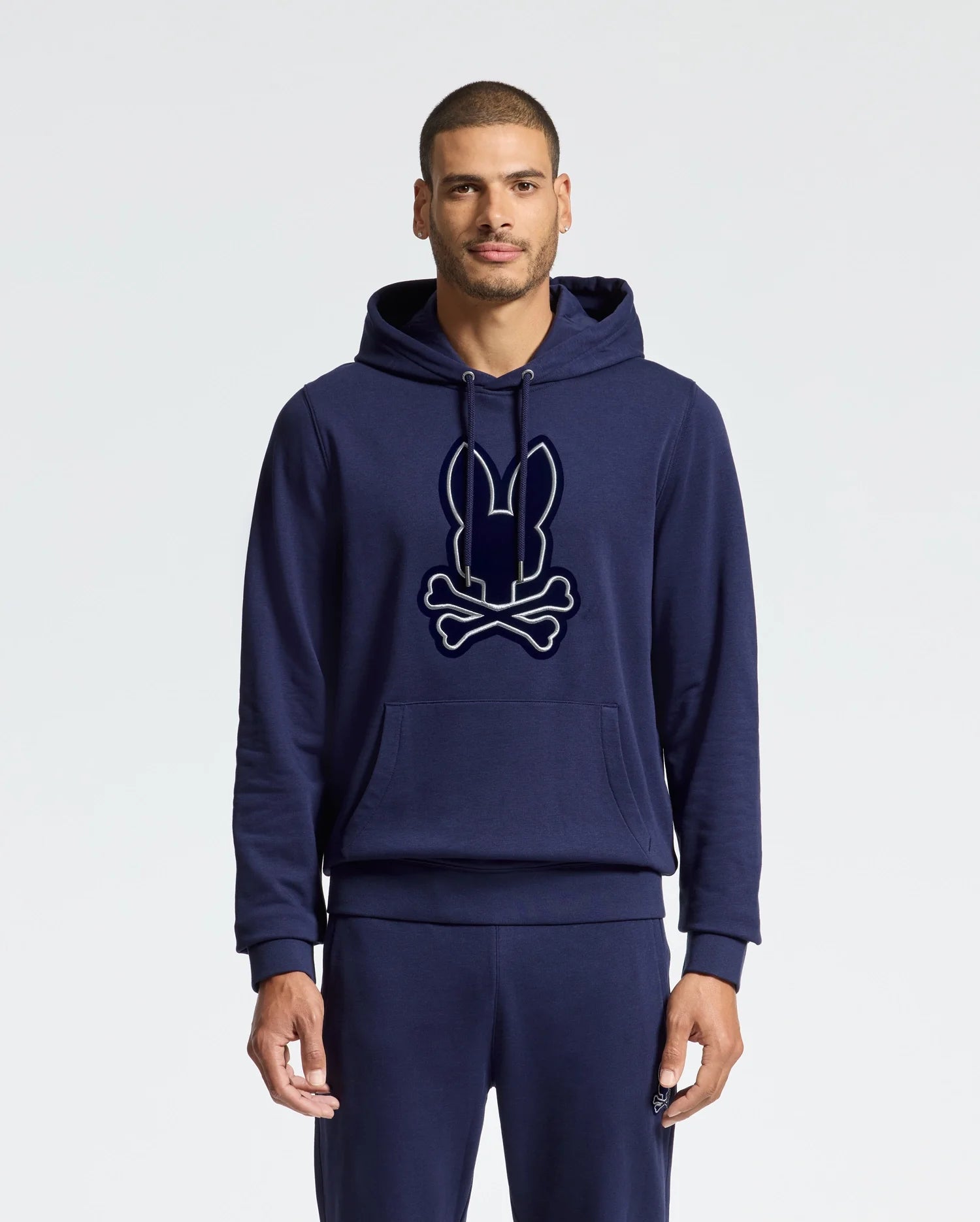 A man is wearing a navy blue MENS ALEXANDER HOODIE - B6H154E200 by Psycho Bunny, adorned with a stylized bunny and crossbones logo on the front. This Everyday Wear Hoodie is crafted from a soft cotton blend and includes convenient features like a kangaroo pocket and drawstrings. The background is plain white.