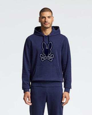 A man is wearing a navy blue MENS ALEXANDER HOODIE - B6H154E200 by Psycho Bunny, adorned with a stylized bunny and crossbones logo on the front. This Everyday Wear Hoodie is crafted from a soft cotton blend and includes convenient features like a kangaroo pocket and drawstrings. The background is plain white.