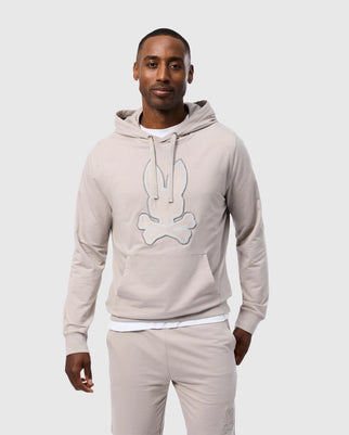 A man is standing and smiling while wearing the comfortable "MENS WALTER LIGHTWEIGHT HOODIE - B6H476D200" by Psycho Bunny, featuring a fuzzy bunny and crossbones design on the front. Made from soft Micro French Terry, it pairs perfectly with his matching beige jogger pants. The background is plain and light-colored.