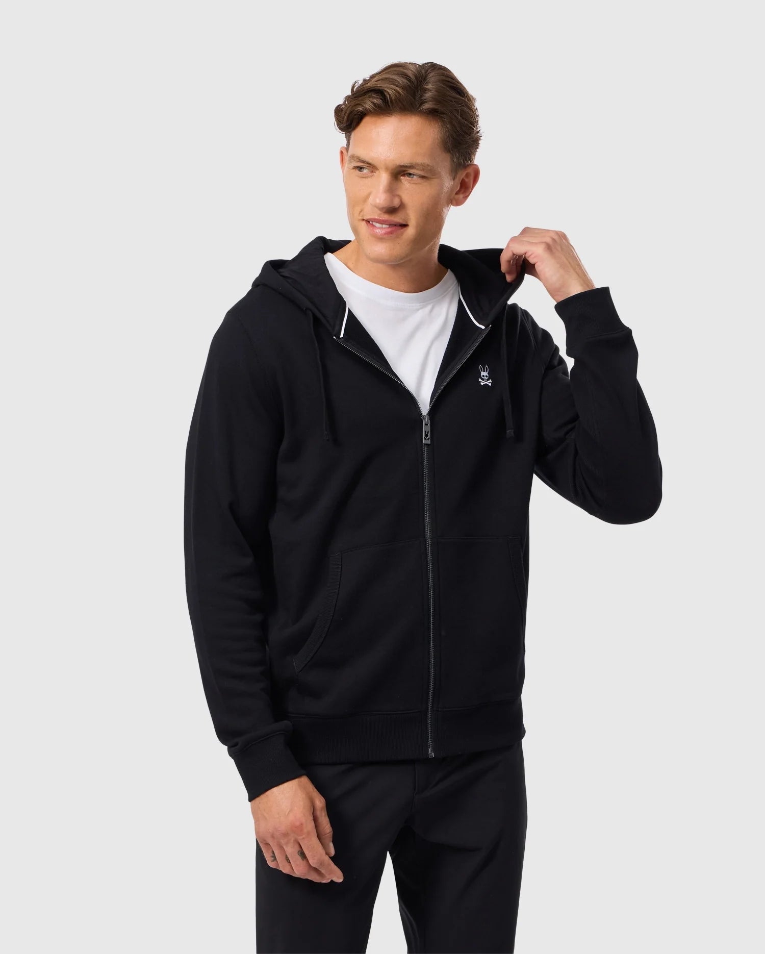 A person wearing a Psycho Bunny MENS LEVY ZIP FLEECE HOODIE (B6H720D200) in black over a white t-shirt stands against a light gray background. The cozy hoodie, crafted from comfortable cotton and featuring a small logo on the chest, frames the person's slight smile as one hand touches the hood.