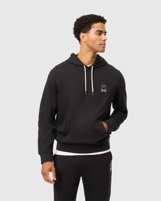 A man with short curly hair is wearing the MENS JAMES PREMIUM HOODIE - B6H789D200 by Psycho Bunny, crafted from essential fleece and adorned with a small embroidered bunny logo on the upper left side along with drawstrings. He pairs it with black pants and stands against a plain white background, facing slightly to the right with his hands by his sides.