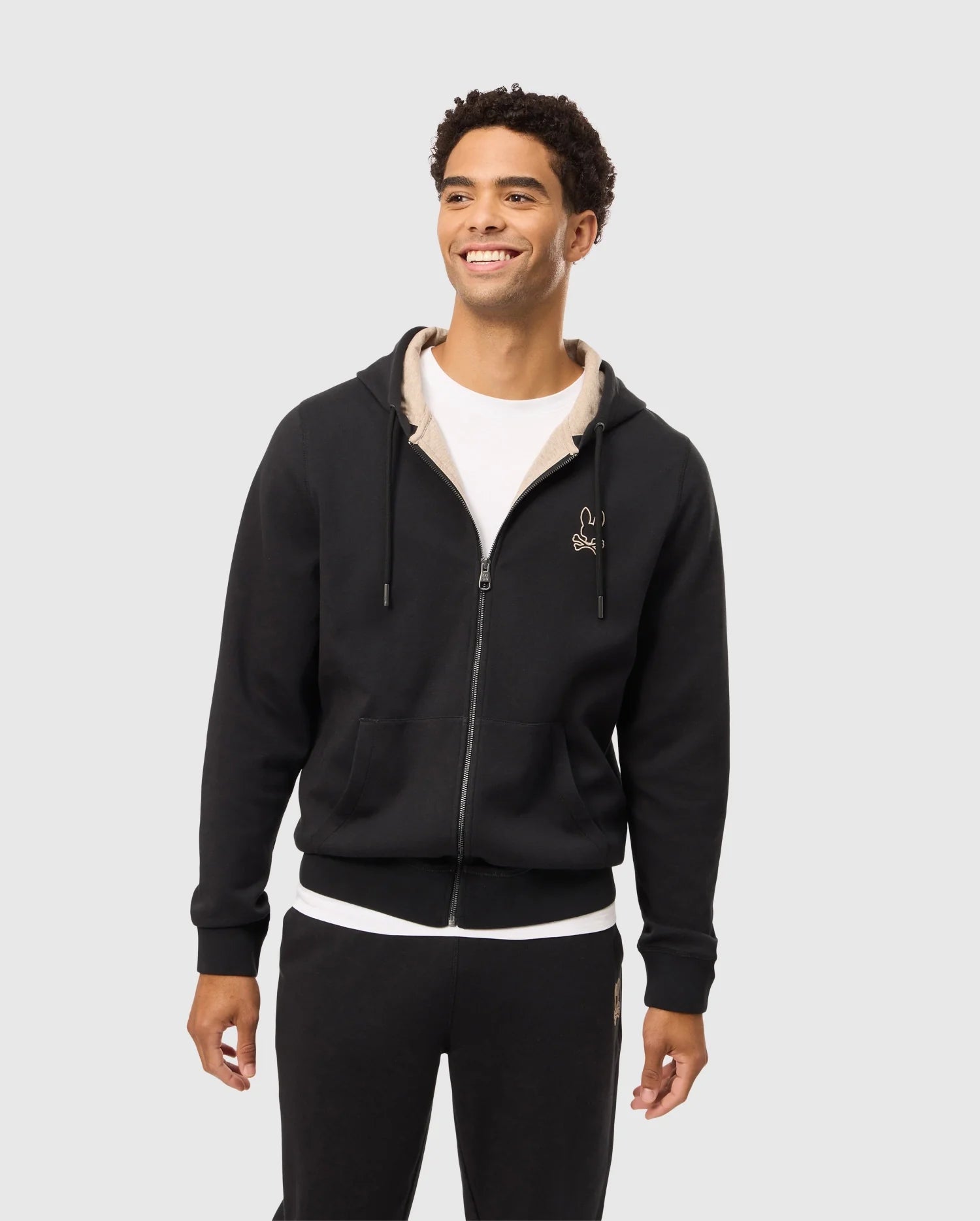 A person smiles while wearing the Psycho Bunny MENS JAMES PREMIUM ZIP HOODIE - B6H793D200, which features an embroidered design on the chest and offers stylish warmth. The black hoodie, with its beige inner lining, is paired with matching black pants. The background is plain and light-colored.