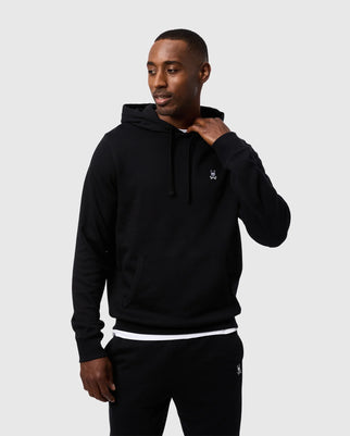 A person with short hair is wearing a black MENS LEVY FLEECE HOODIE - B6H859D200 by Psycho Bunny, featuring a small white logo on the left side of the chest. They are standing against a plain white background, looking to their left and casually holding their neck with their right hand.