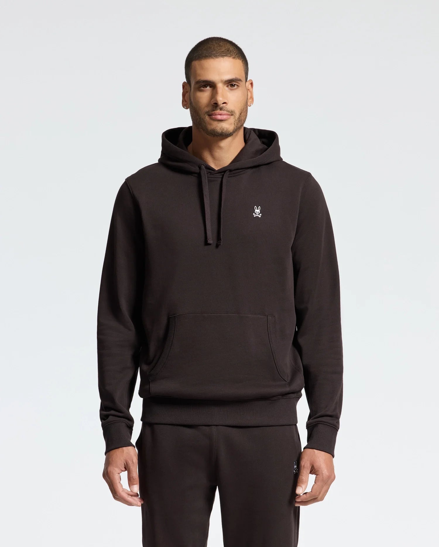 A person in a Psycho Bunny MENS LEVY ESSENTIAL FLEECE HOODIE - B6H859D200, crafted from 100% cotton, faces the camera. The black hoodie features a small chest logo and matches their regular-fit black pants. They have short hair and a beard against a plain, light-colored background.