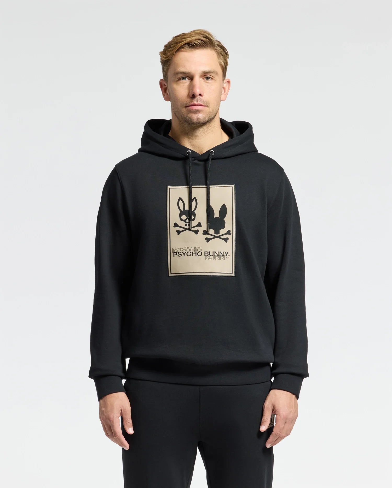 A man stands wearing the black Mens Monroe Twill Patch Fleece Hoodie, featuring a tan rectangle with two bunny logos and 