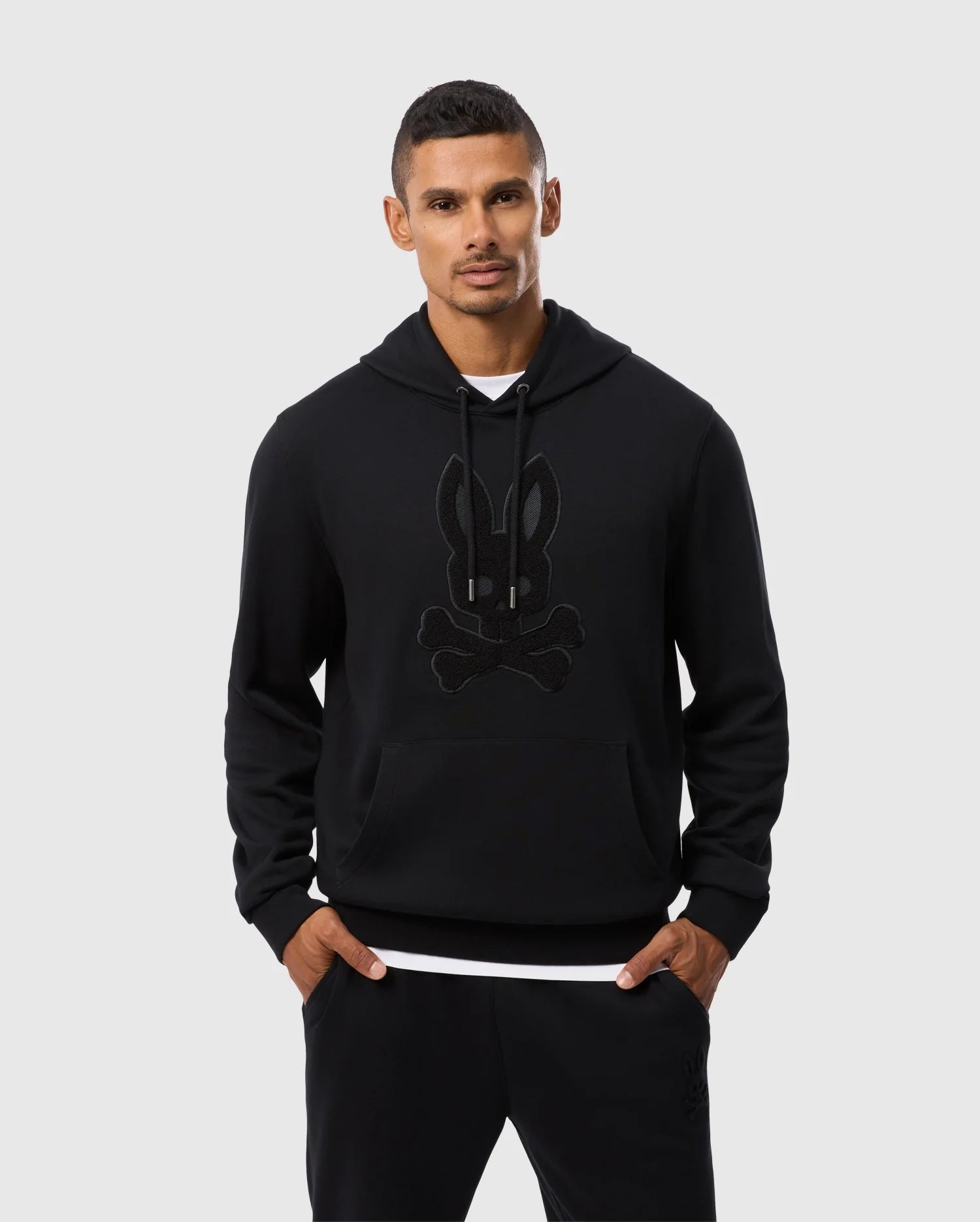 A man wearing a Psycho Bunny MENS PACIFIC CHENILLE HOODIE - B6H930D200, which features a black French terry fabric with a bunny and crossbones chenille embroidery patch, stands against a plain background. He has short dark hair, is clean-shaven, and has his hands in the hoodie pockets. He is also wearing black pants and a white shirt that is partially visible.