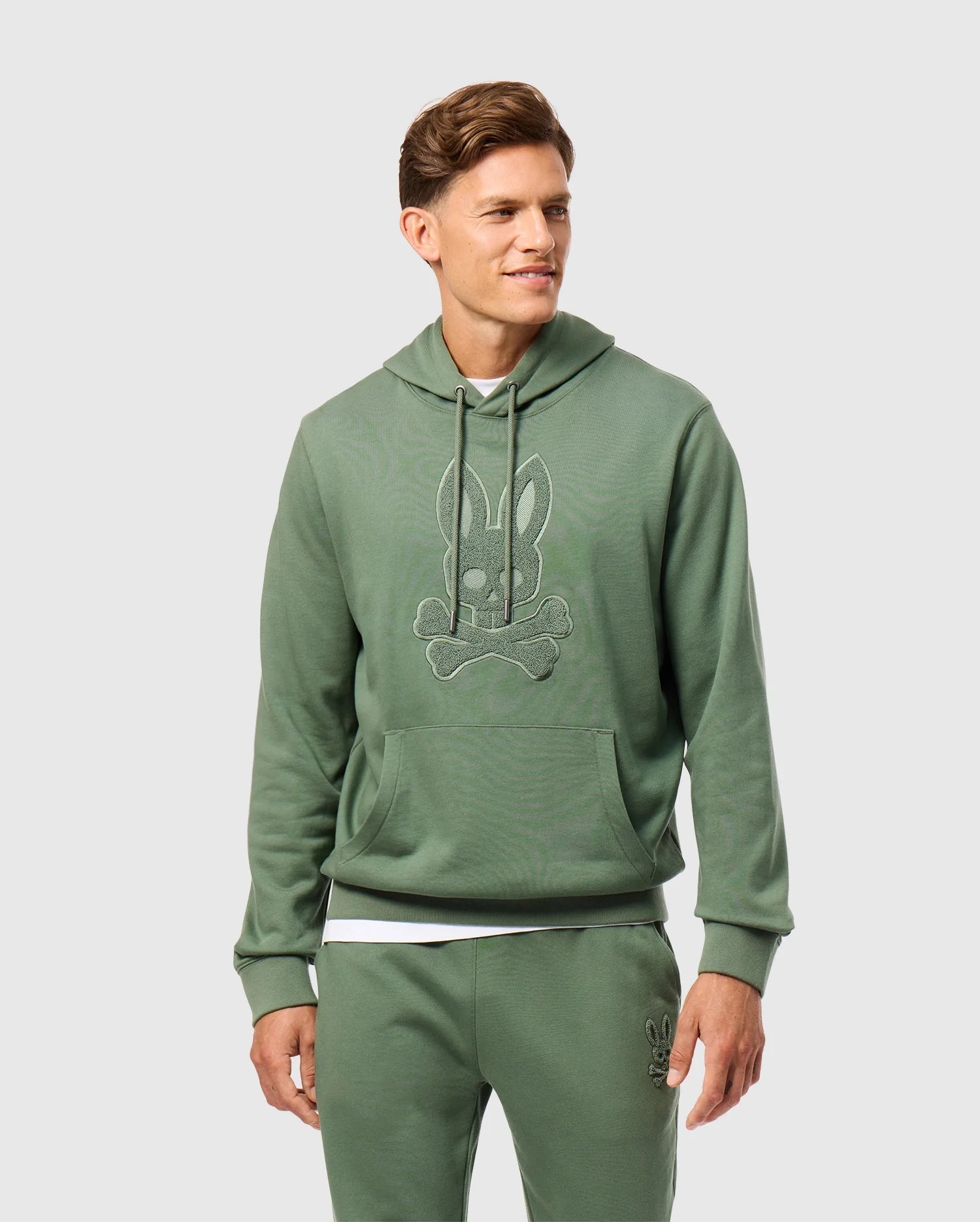 A person is wearing the Psycho Bunny MENS PACIFIC CHENILLE HOODIE in green, decorated with a rabbit logo made from chenille embroidery, along with matching pants. The hoodie showcases a design of a rabbit head above crossed bones. The person is smiling and gazing to the side against a light-colored background.