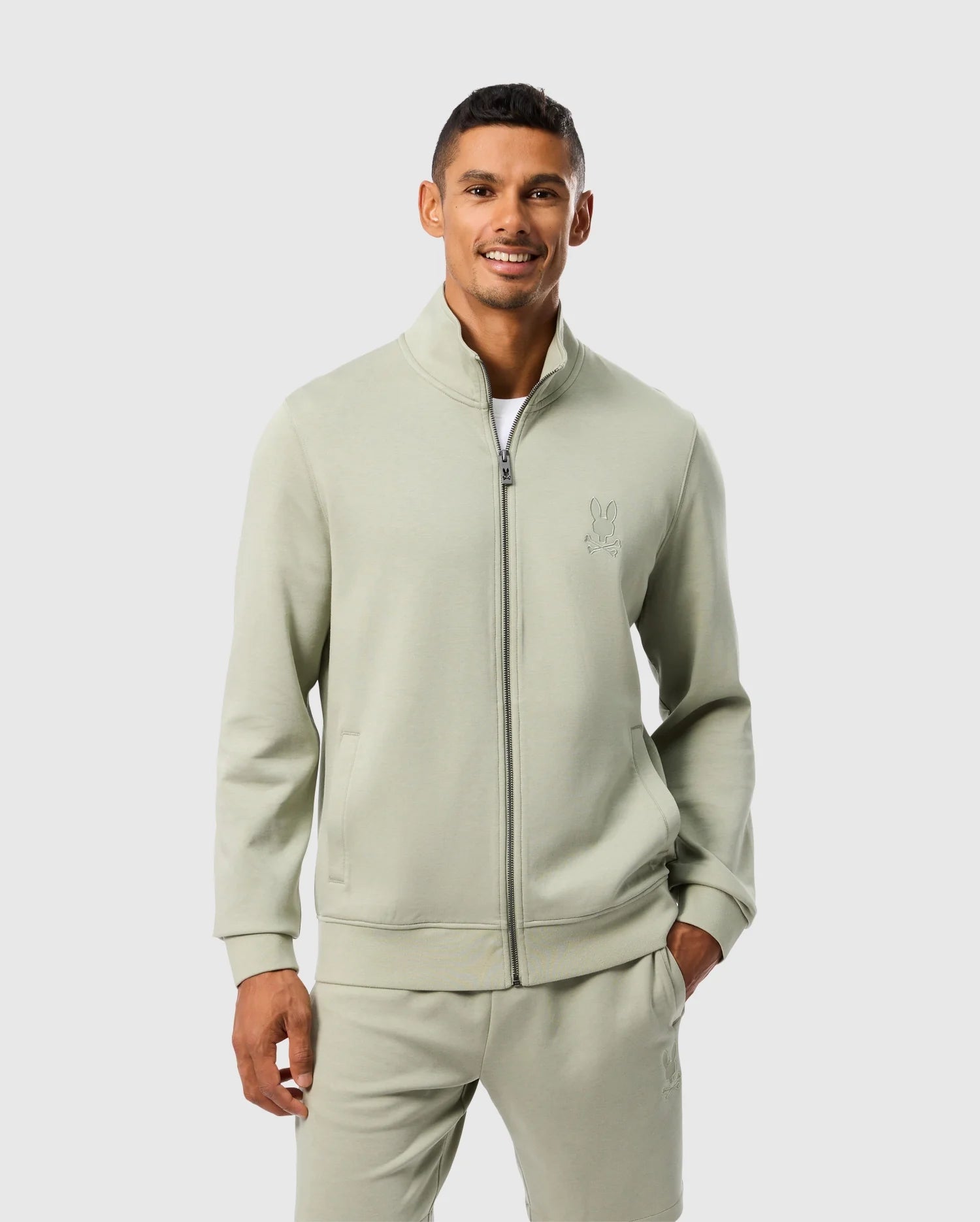 A man wearing a Psycho Bunny MENS LYONS HIGH COLLAR FULL ZIP - B6J380B200 track-style sweatshirt and matching pants stands against a plain white background. He is smiling and has his hands in his pockets. The high-collar jacket features a small embroidered logo on the left chest.