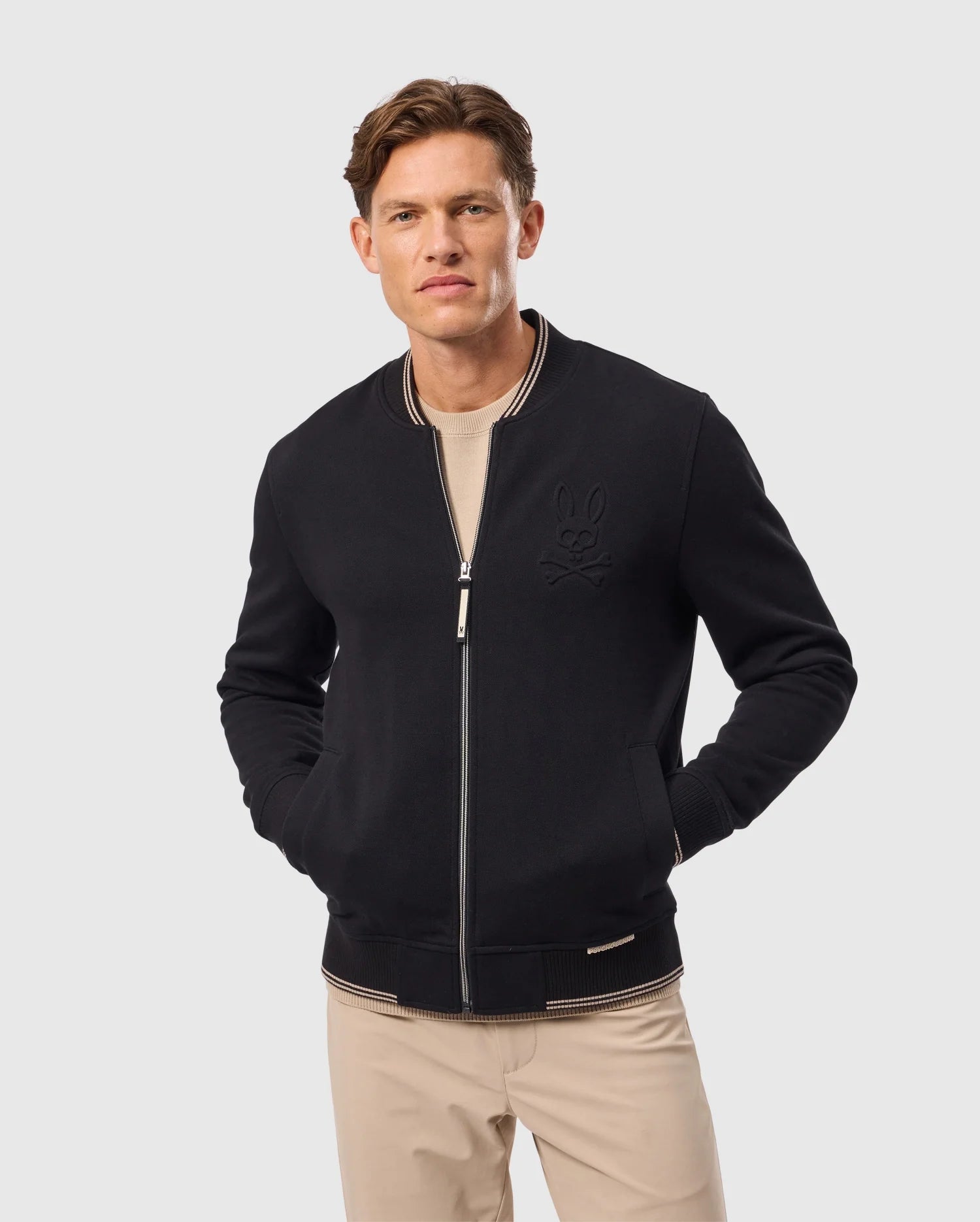 Wearing a Psycho Bunny MENS BRENT PIQUE KNIT BOMBER JACKET - B6J743D200, a man stands against a plain light background. The black bomber jacket features a subtle embossed logo on the chest, complemented by a beige shirt underneath and khaki pants, with his hands in the jacket pockets.