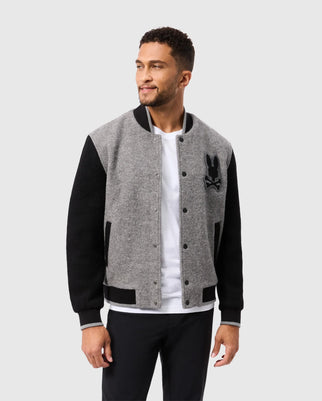 A man with short hair and a beard is wearing a Psycho Bunny MENS TOM WOOL BOMBER JACKET - B6J744D200 with a rabbit logo on the left chest, over a white t-shirt. He stands looking to his left with a neutral expression and has his hands at his sides. The background is plain gray.