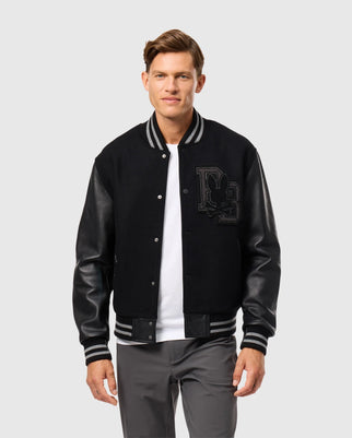 A person wearing the Psycho Bunny Men's Brayden Wool Leather Varsity Bomber, featuring leather sleeves and the embroidered initials "DG" on the left side, paired with a white shirt. They are standing against a plain gray background.