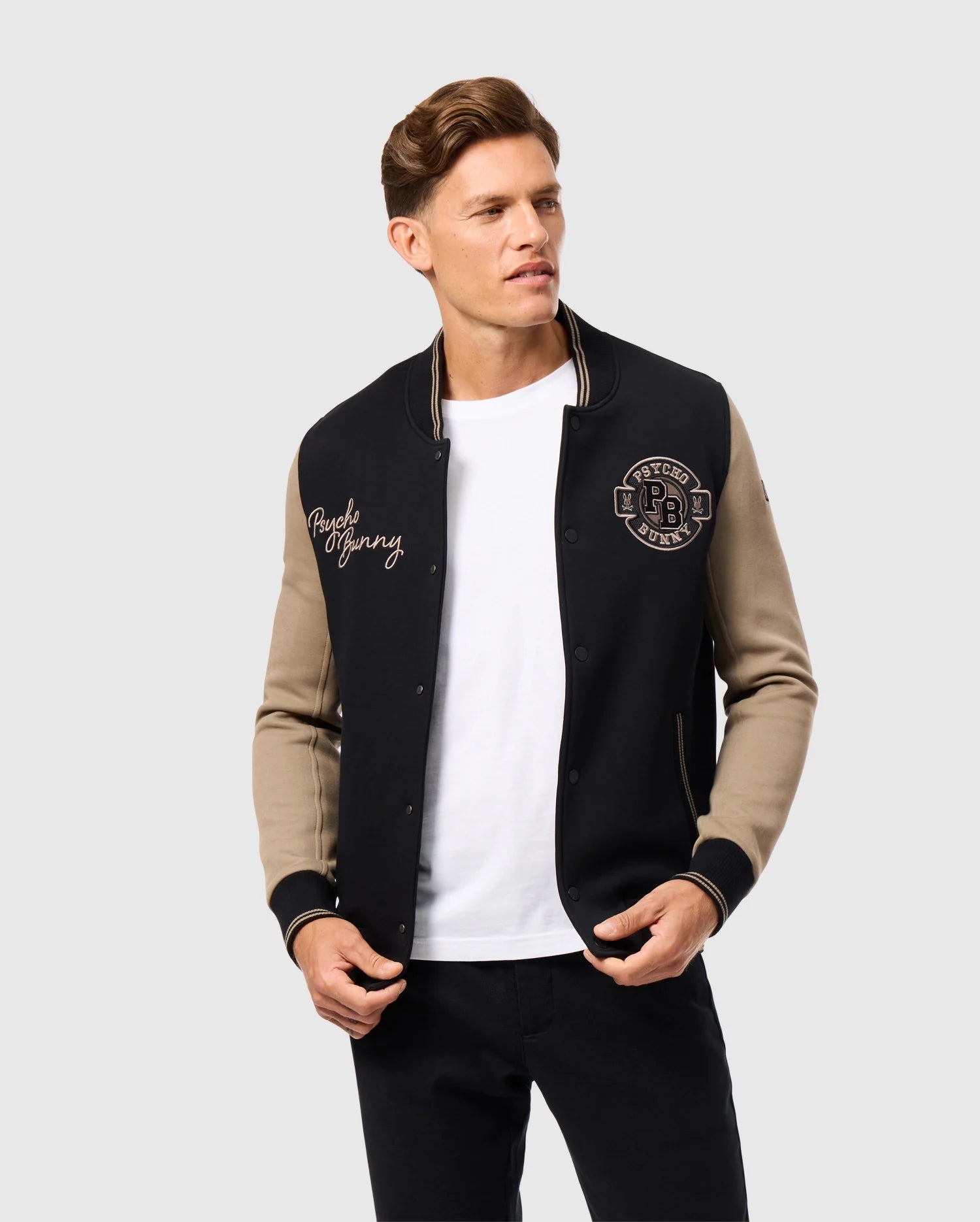 A person with styled brown hair is wearing the Psycho Bunny MENS ZACK VARSITY JACKET - B6J878D200, a black varsity jacket made from sandwich jersey fabric, featuring beige sleeves and embroidered patches on the chest. Dressed in a white T-shirt and black pants, they stand against a plain light grey background.