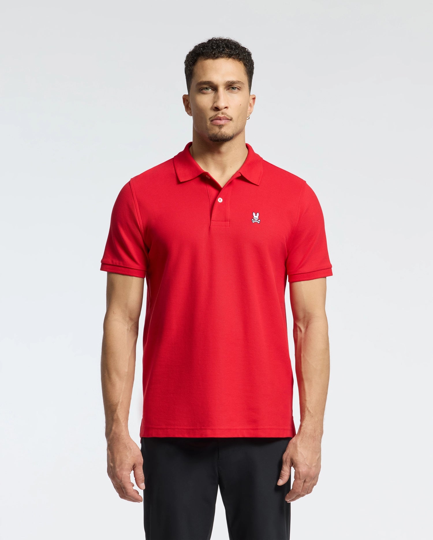 A man in a Psycho Bunny MENS CLASSIC PIQUE POLO SHIRT - B6K001CRPC, made from Pima cotton with mother-of-pearl buttons, stands against a plain background. The red shirt has a small embroidered logo on the left chest. His short hair complements his direct gaze at the camera.