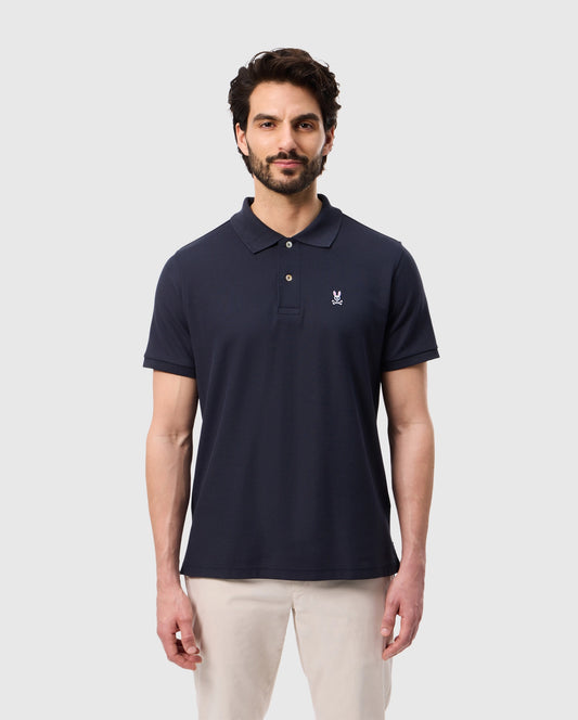 A man with a beard is wearing Psycho Bunny's men's classic pique polo shirt in navy, made from 100% Pima cotton. It features mother-of-pearl buttons and a discreet embroidered logo on the left chest. He stylishly pairs it with light-colored pants against a plain light gray background.