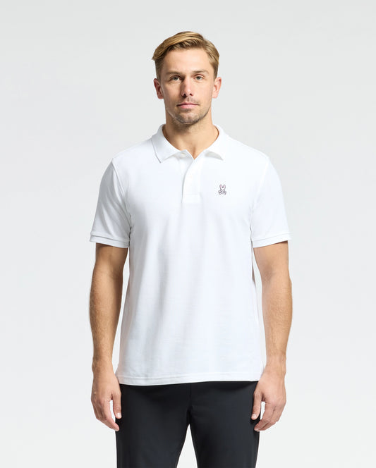 A man wearing a Psycho Bunny MENS CLASSIC PIQUE POLO SHIRT, identified by the small logo on the chest, stands against a plain background. He has short blond hair and is dressed in dark pants.