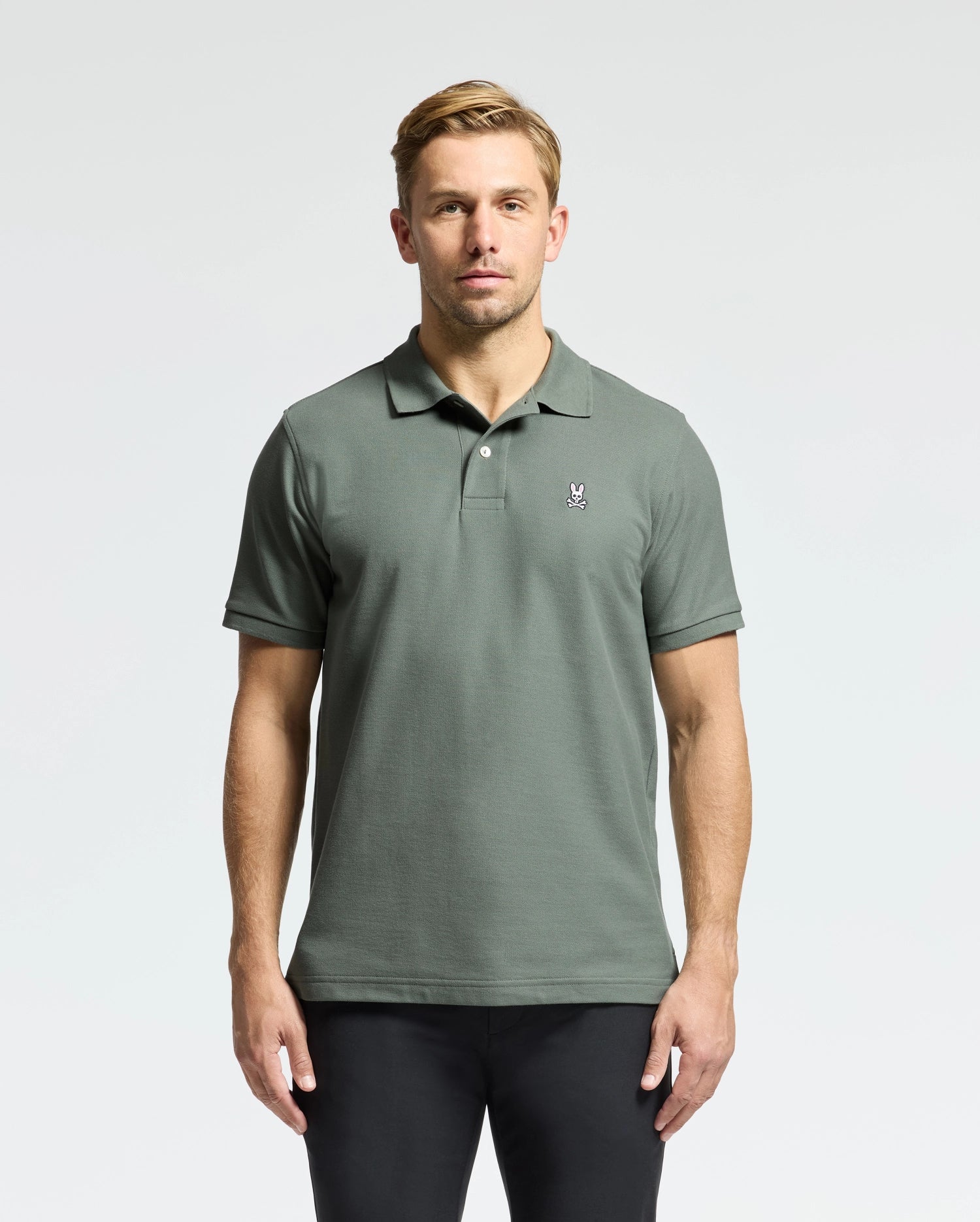 A man in a green Psycho Bunny Men's Classic Piqué Polo (B6K001B200) with an embroidered Bunny logo stands against a white background. He has short, light brown hair and wears black pants made from soft Pima cotton.