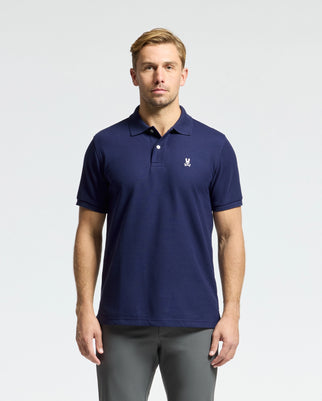 A person wearing a Psycho Bunny MENS CLASSIC PIQUE POLO SHIRT - B6K001CRPC in navy blue, featuring a regular fit and crafted from soft pima cotton, with a small white logo on the chest, stands against a plain white background. They have short blond hair and are looking directly at the camera.