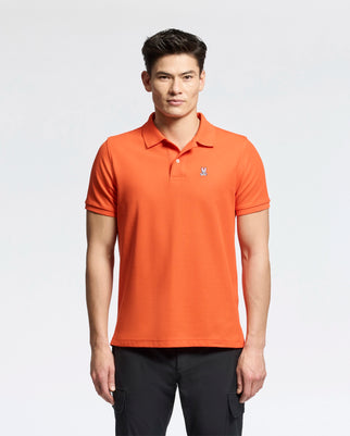 A person models the Psycho Bunny Men's Classic Piqué Polo Shirt in orange, featuring a small logo, against a plain white background. They pair it with dark pants and have short, styled hair.