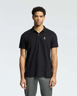 A person stands against a plain background wearing Psycho Bunny's MENS ALEXANDER PIQUE POLO - B6K101E200, a black men's Pima cotton polo with an embroidered chest design. They have short curly dark hair and are paired with gray pants.