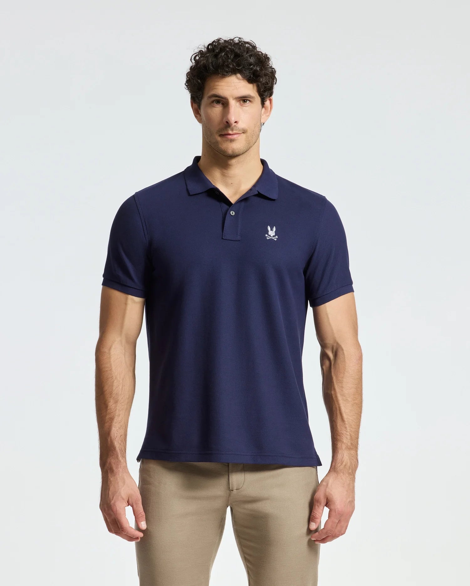 A man with short curly hair wears beige pants and a navy blue Psycho Bunny MENS ALEXANDER PIQUE POLO - B6K101E200, featuring a small white emblem on the chest. Made from four-way stretch Pima cotton, this polo offers durable breathability as he stands against a plain white background.