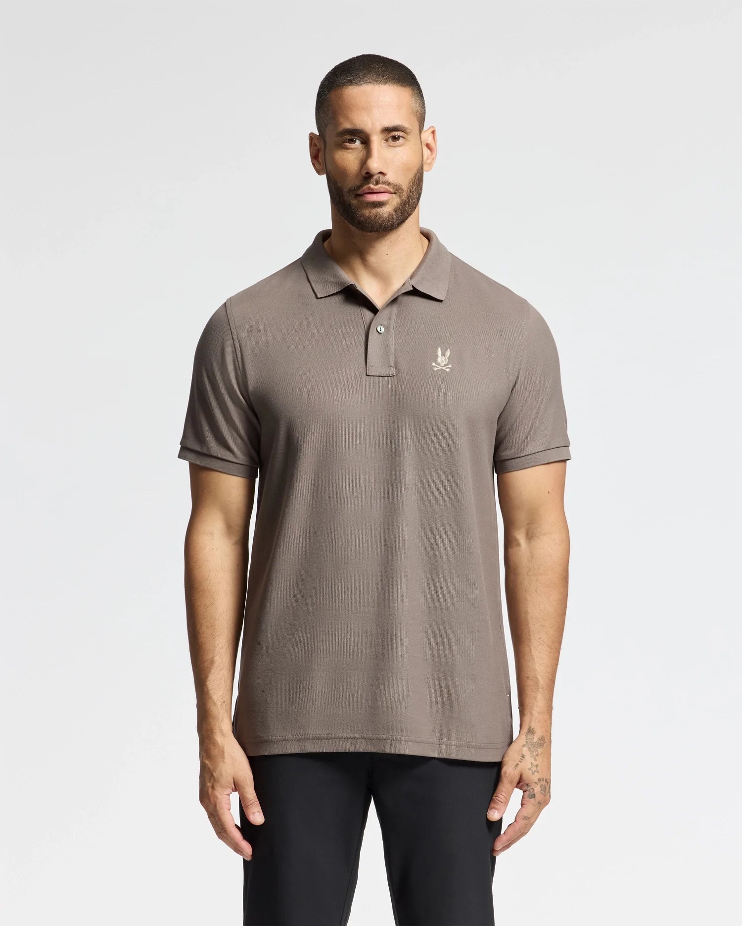 A person wearing the Psycho Bunny MENS ALEXANDER PIQUE POLO - B6K101E200, featuring a small embroidered logo on the chest, stands against a plain background. The lightweight design of the polo complements their short hair and beard as they gaze toward the camera, hands relaxed at their sides.