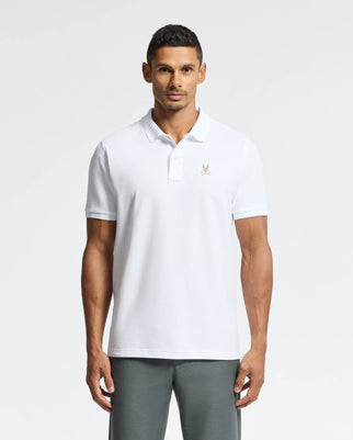 A man wearing a white MENS ALEXANDER PIQUE POLO - B6K101E200 by Psycho Bunny, featuring an eagle logo on the chest, stands against a plain background. With short dark hair, he gazes directly at the camera, highlighting the elegance and comfort of this lightweight men's polo shirt.