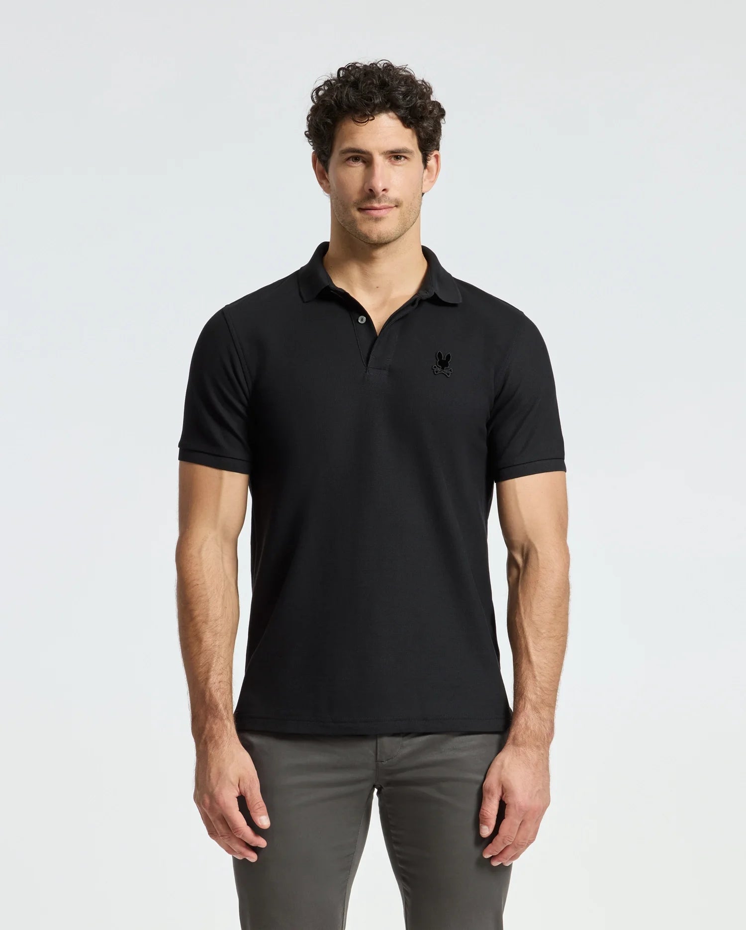 A person wearing a black Men's Branson Pique Polo - B6K108E200 from Psycho Bunny and gray pants stands against a plain white background.