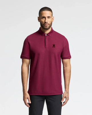 A man with short hair and a beard is wearing the Psycho Bunny MENS BRANSON PIQUE POLO - B6K108E200 in burgundy, which features a small black velvet patch logo on the chest. He pairs it with black pants and stands against a plain light background.