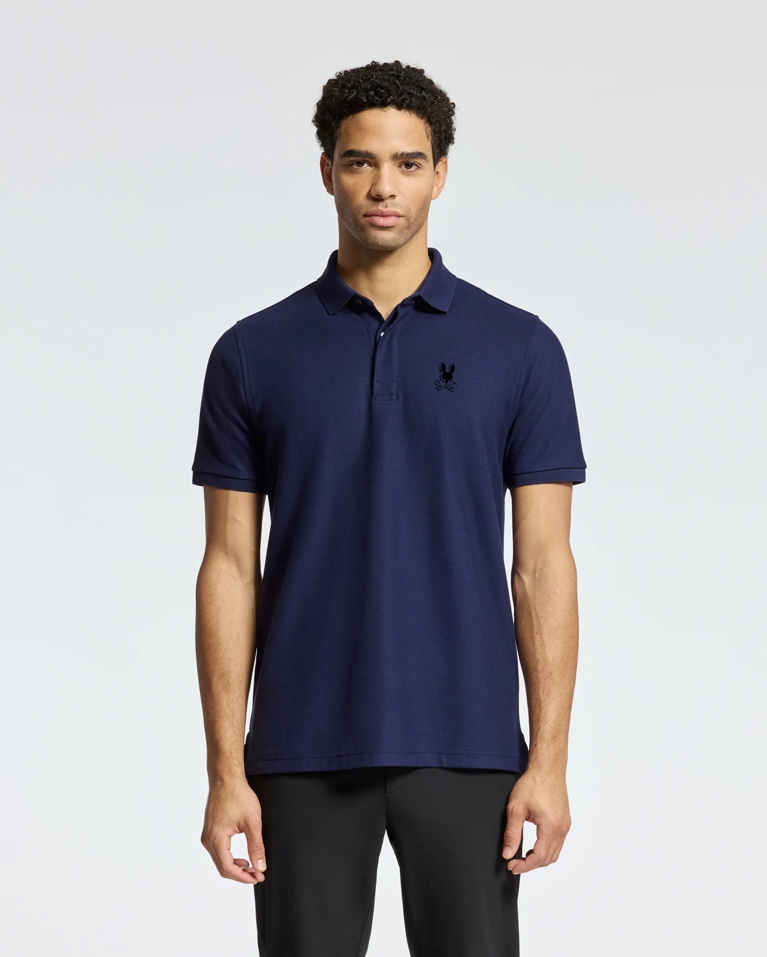 Against a plain light background, a person with short curly hair wears black pants and the Psycho Bunny Men's Branson Pique Polo (B6K108E200) in luxurious dark blue 100% Pima cotton, featuring a small embroidered logo on the left chest.