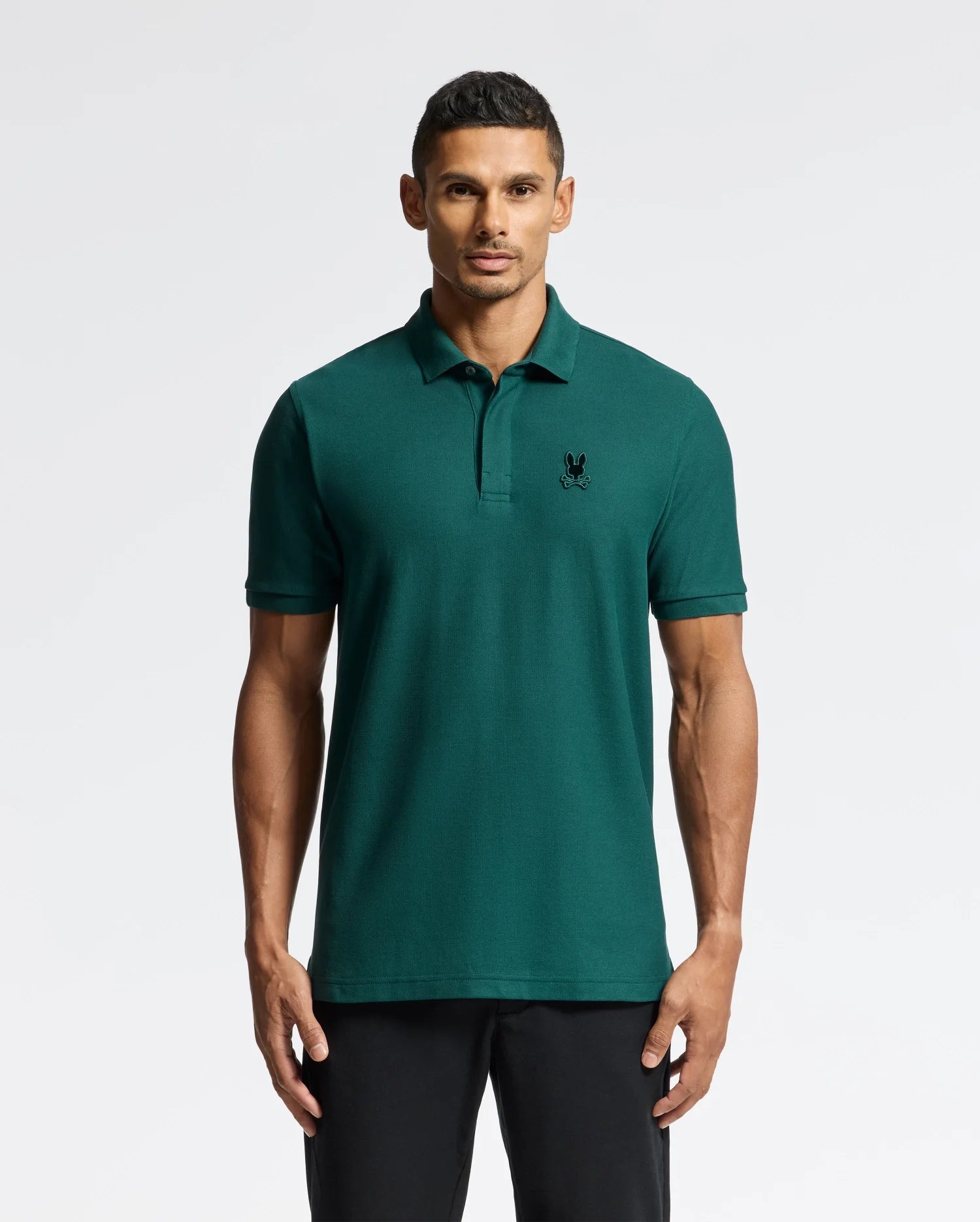 A person wearing a luxurious dark green Psycho Bunny Men's Branson Pique Polo (B6K108E200), made from 100% Pima Cotton, features a subtle logo on the left chest as they stand against a plain light gray background. With short hair, they gaze directly at the camera.