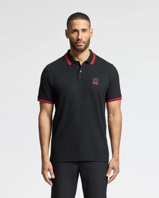 A man with a buzz cut is wearing the Psycho Bunny MENS PIERCE PIQUE POLO - B6K201E200, featuring black Pima cotton with red trim and an embroidered Bunny emblem on the chest. He's standing against a plain white background, looking straight ahead with his arms relaxed by his sides.