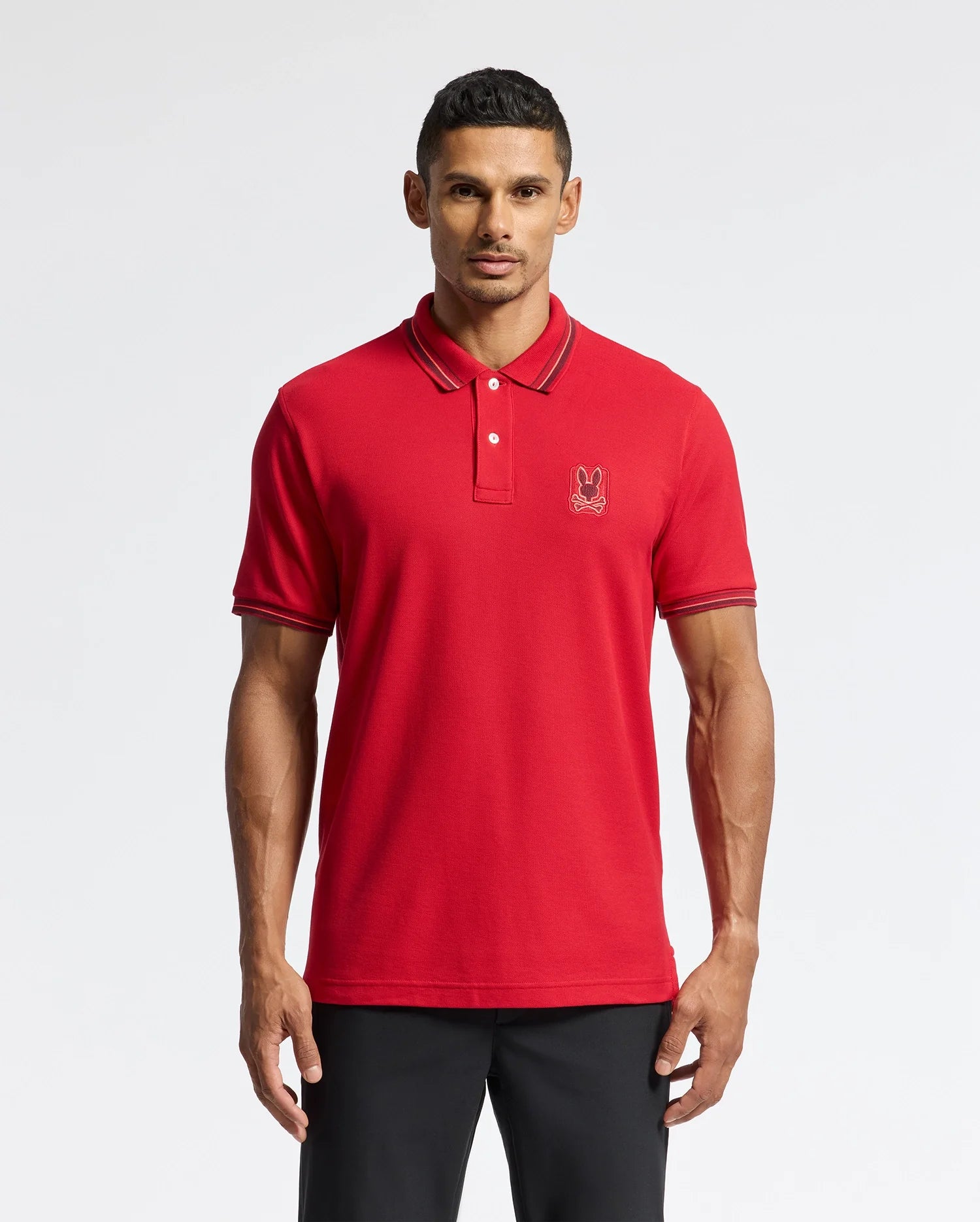 A person wearing a red Psycho Bunny MENS PIERCE PIQUE POLO - B6K201E200, featuring an embroidered bunny emblem on the left side of the chest, paired with black pants, stands against a plain white background.