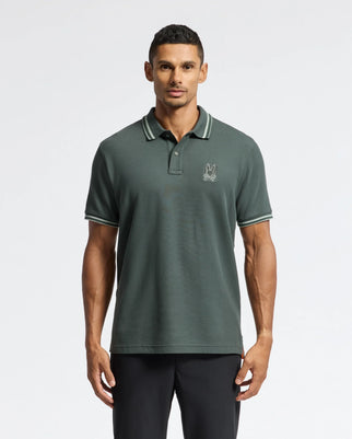 A man stands against a plain background wearing a dark green Psycho Bunny MENS PIERCE PIQUE POLO - B6K201E200. The polo shirt features Mother-of-Pearl buttons and an embroidered Bunny logo on the chest, complete with a collar and short sleeves. It is stylishly paired with sleek black pants.