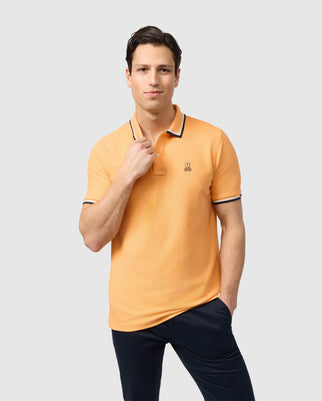 A person with short dark hair is wearing a light orange Psycho Bunny MENS KINGSBURY PIQUE POLO SHIRT - B6K235B200 with a small embroidered logo on the chest. They are posing with one hand touching their collar and the other in their pocket. The background is plain and light gray, highlighting the classic silhouette.