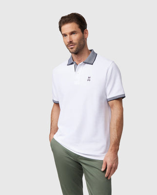 A man wearing a Psycho Bunny Men's Southport Pique Polo Shirt (B6K263B200) featuring a contrasting dark collar and sleeve trim stands casually with one hand in his pocket. He pairs it with light olive-green pants, and the Pima cotton polo perfectly complements the plain light gray background.