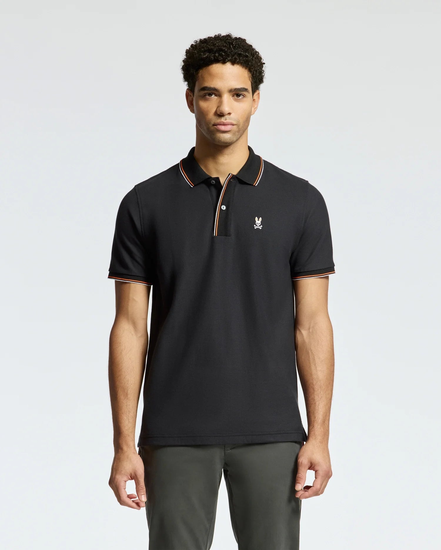 A man wearing the Psycho Bunny MENS BRANDON PIQUE POLO - B6K280E200 with orange trim and a small logo stands against a plain background. The luxurious design accentuates his short, curly hair as he looks straight at the camera.