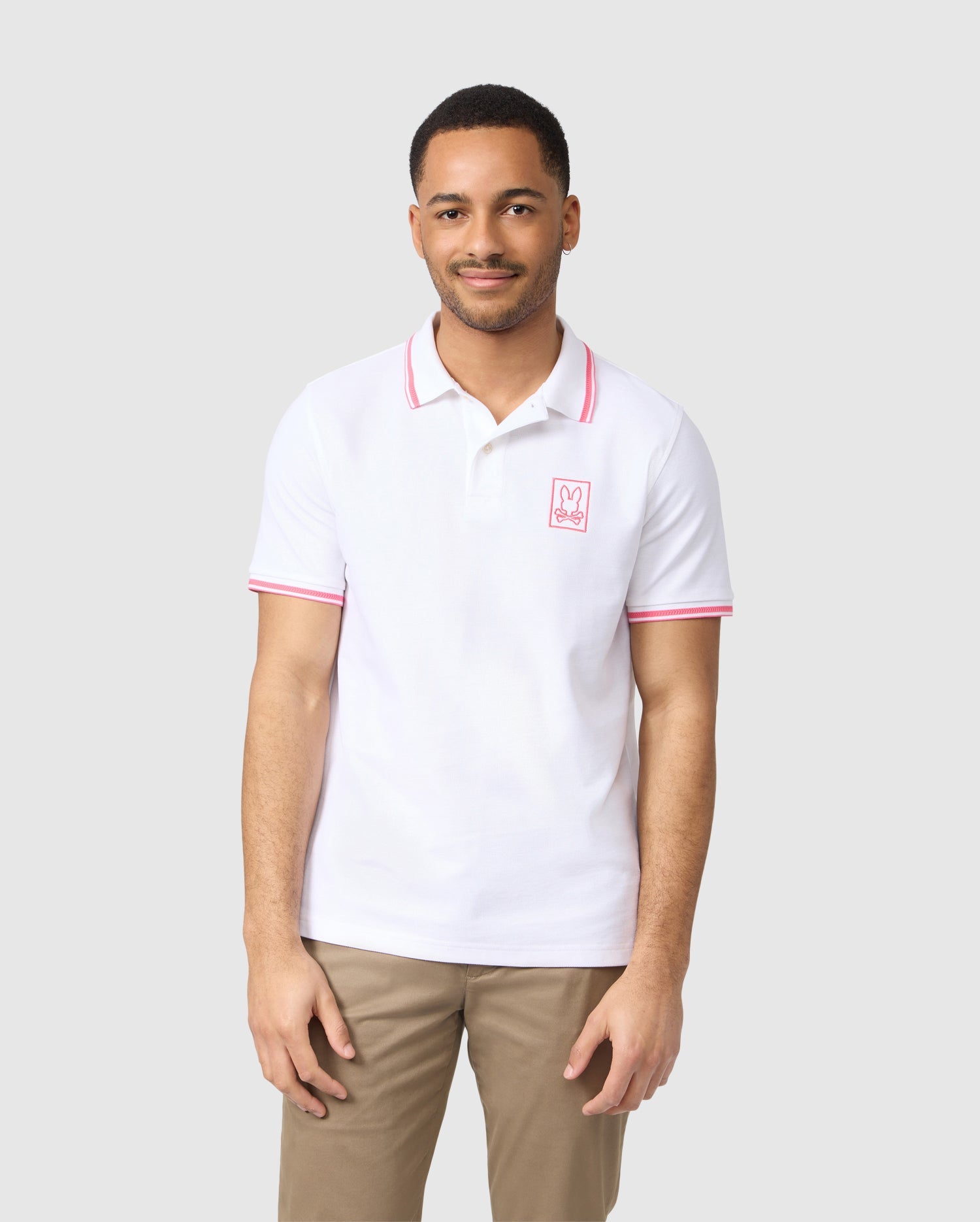 A man is standing against a plain white background, wearing a Psycho Bunny MENS ARCADIA PIQUE POLO SHIRT - B6K406B200 made from soft Peruvian Pima cotton. The shirt features red trim on the collar and sleeves and a small red bunny logo on the left side of the chest. He has short hair and a trimmed beard, and he is looking at the camera while smiling.