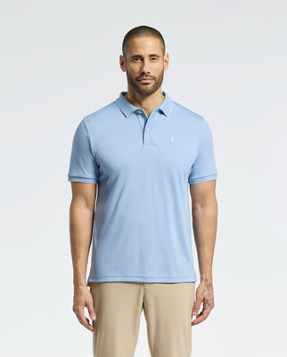 A bearded man in a Psycho Bunny Mens Bryce Performance Pique Polo (B6K486F200), light blue, and khaki pants stands against a plain white background. The Quick Dry fabric enhances his right arm tattoos, providing style and breathability.