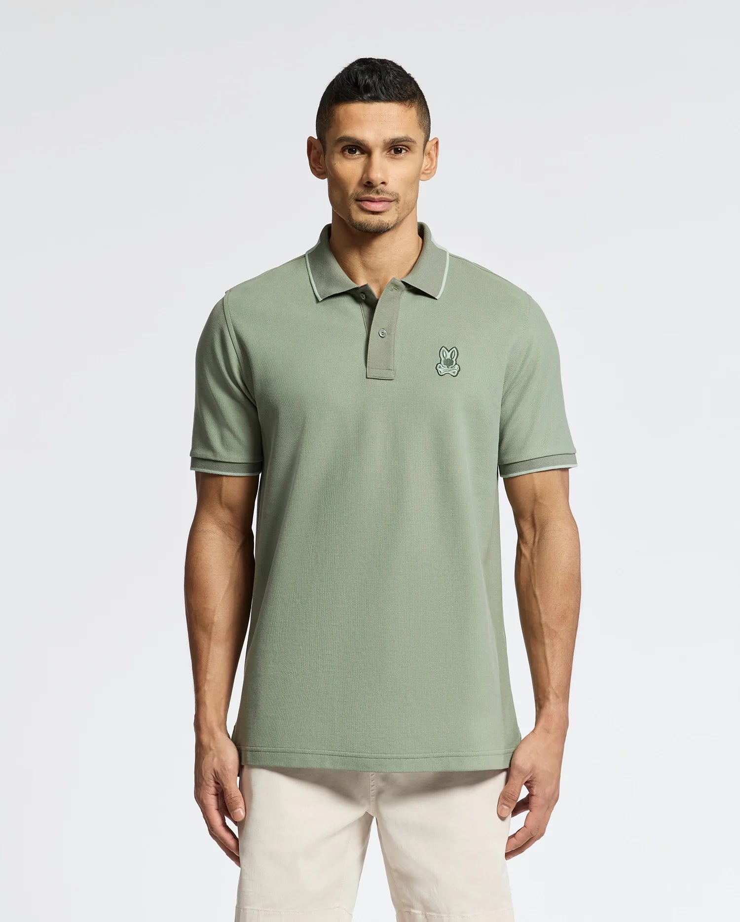 A man wears the Psycho Bunny MENS NEW CASTLE PIQUE POLO - B6K487F200 in light green, with a small rabbit logo on the chest, luxury finishes including a collar and short sleeves, paired with beige pants against a plain white background.