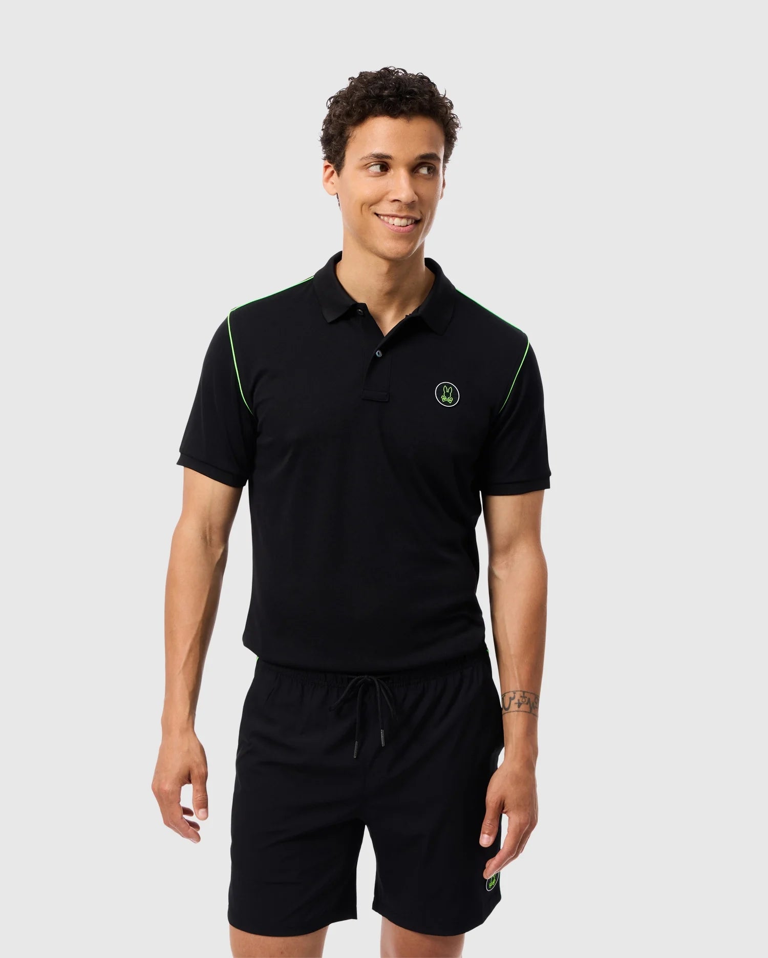 A man wearing a black Psycho Bunny MENS WEBSTER SPORT POLO SHIRT - B6K580C200, which features moisture-wicking material, is paired with black shorts. The outfit includes green accents and a small logo on the left chest, making it ideal for staying cool while he stands and smiles against a plain light gray background.