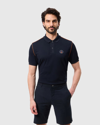 A man with dark hair and a beard is wearing the Psycho Bunny MENS WEBSTER SPORT POLO SHIRT - B6K580C200, a navy blue polo featuring orange piping on the shoulders and a small emblem on the chest. The breathable fabric pairs perfectly with his matching navy blue shorts as he stands with one hand in his pocket against a plain light-gray background.