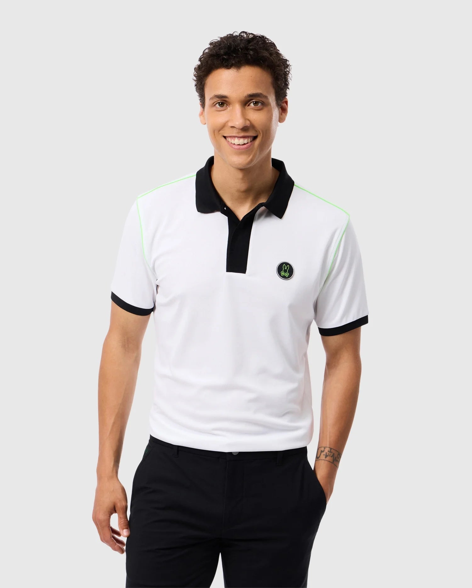 A smiling man with short curly hair is wearing the Psycho Bunny MENS WEBSTER SPORT POLO SHIRT - B6K580C200, a white polo featuring black collar and sleeve trim along with a small green and black logo on the chest. Made from moisture-wicking fabric for a comfortable fit, he pairs it with black pants while standing against a plain light gray background.