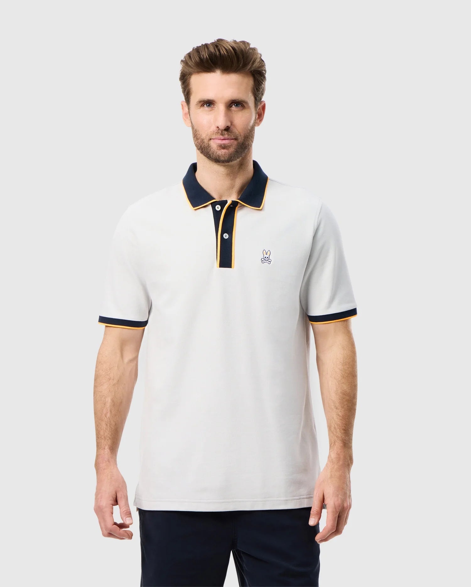 A man with short hair and a beard is wearing an elegant Psycho Bunny MENS DALLAS PIQUE POLO SHIRT - B6K602C200. The white polo features a black collar, black trim on the sleeves, two buttons, and a small emblem embroidered on the left chest. He stands against a plain, light-colored background.