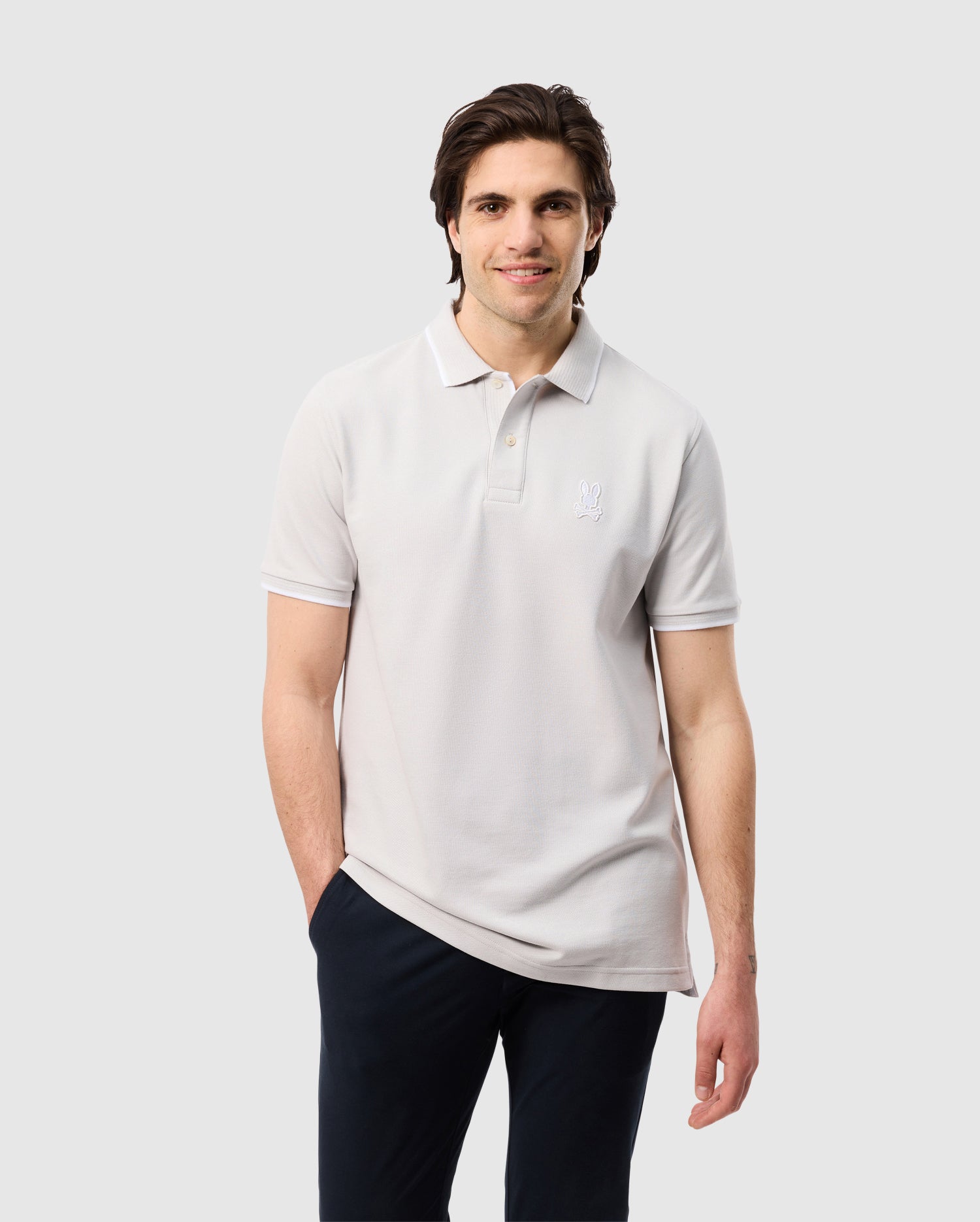 A man with dark hair is smiling and wearing a light gray Psycho Bunny MENS HOUSTON PIQUE POLO SHIRT - B6K604C200 with mother-of-pearl buttons and an embroidered Bunny design on the chest. He has one hand in his pocket and is standing against a plain white background, paired with dark pants.